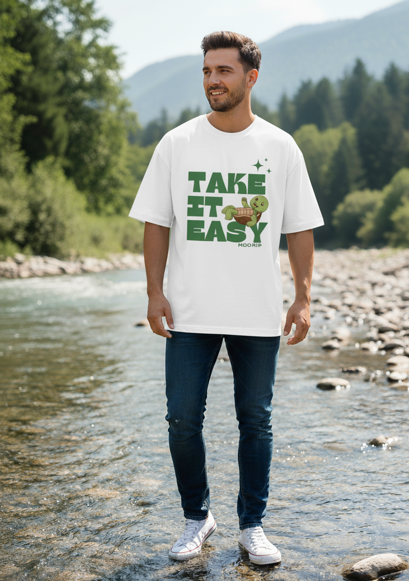 Man wearing a white t-shirt with 'Take It Easy' text standing by a river with mountains in the background