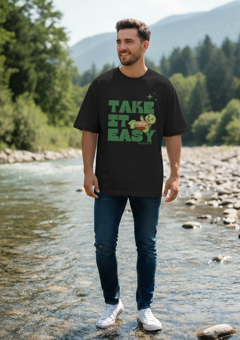 Man wearing a black t-shirt with 'Take It Easy' text standing by a river with mountains in the background